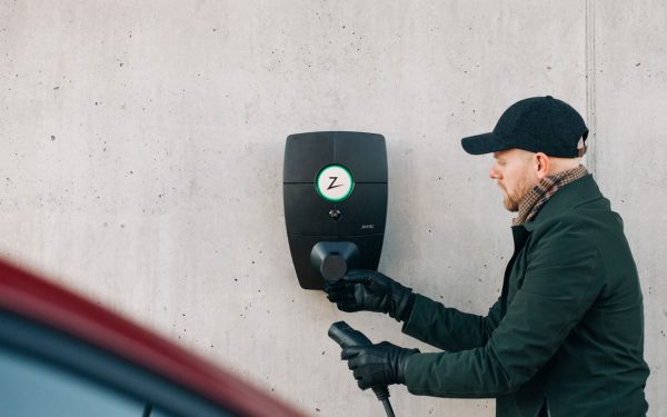 mann med mørk jakke og caps bruker en sort, moderne elbillader montert på grå betongvegg.